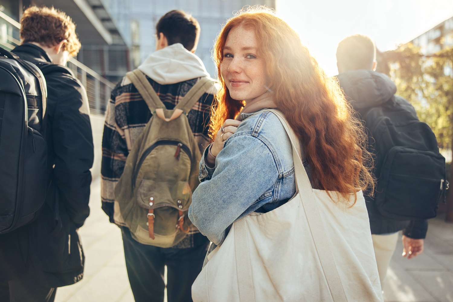 Studentin mit roten Haaren, blickt in die Kamera. Im Hintergrund mehrere Personen und ein Sonnenstrahl.